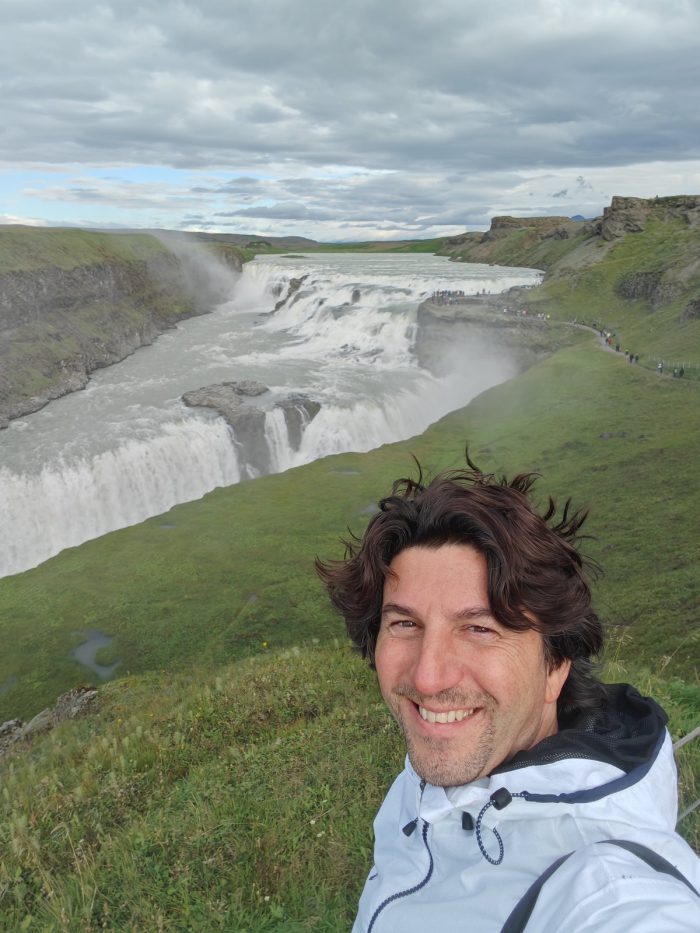 Selfie davanti a una grande cascata del Golden Circle, con il salto d'acqua e il bordo erboso del canyon sullo sfondo